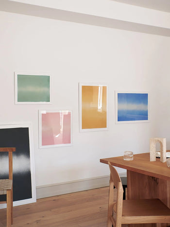 A minimal dining space with a wooden table, chairs, clear glasses, and Anne Nowak - Yellow Interstellar (30x40) by The Poster Club hanging on a white wall.