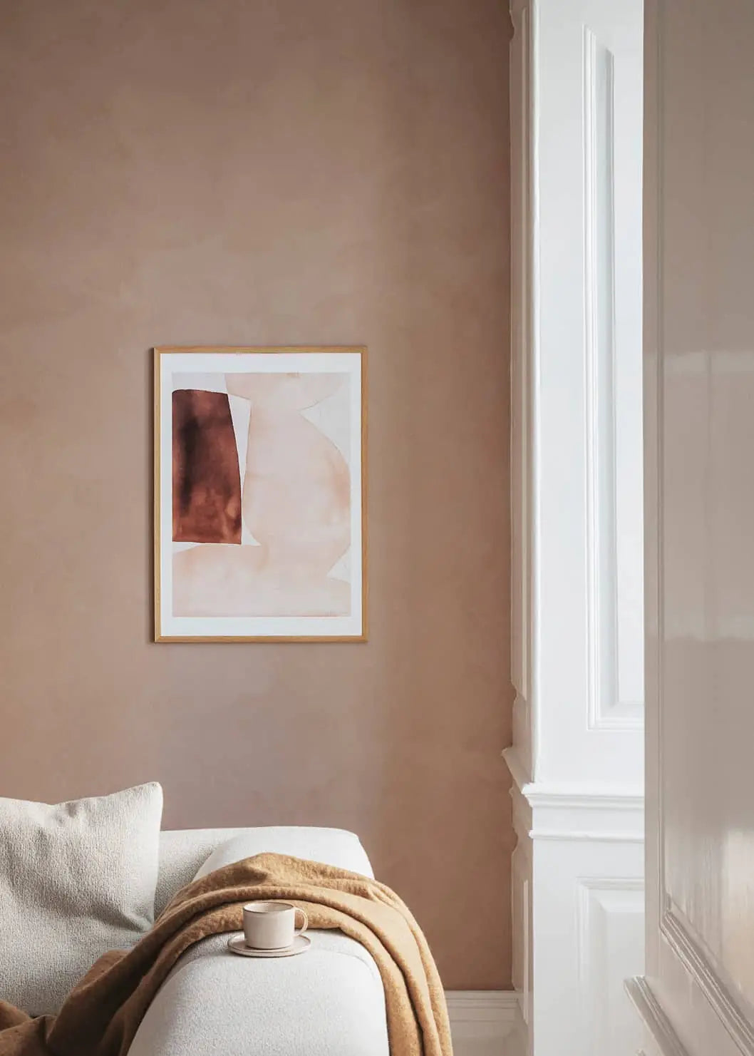 A minimalist living room features a beige wall with The Poster Club Berit Mogensen Lopez - Improvise (50x70) art print in a wooden frame, a white sofa with a tan blanket, and a coffee cup near a large window with natural light.