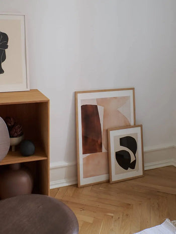 The Poster Club’s Berit Mogensen Lopez Improvise (50x70) art print in a minimalist wooden frame leans against a white wall, beside a wooden shelf with decor and a round brown seat in the foreground.