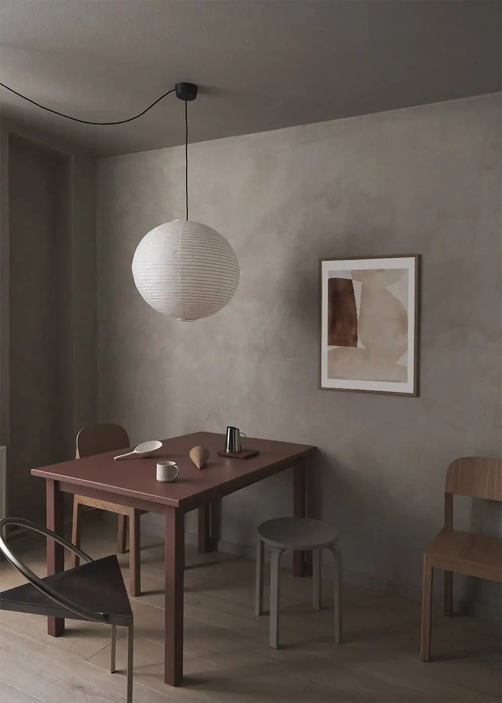 A minimalist dining area with a reddish-brown table, assorted chairs, ceramic mugs, a metal pitcher, paper lantern pendant light, and The Poster Clubs Berit Mogensen Lopez - Improvise (50x70) art print on a gray textured wall above light wood flooring.