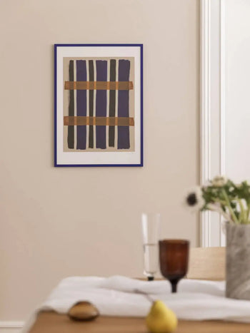 The Poster Club Berit Mogensen Lopez - Checkered (50x70) print, with abstract blue and brown lines, hangs on a beige wall above a table arranged with a glass, dark goblet, flowers, and a yellow pear.