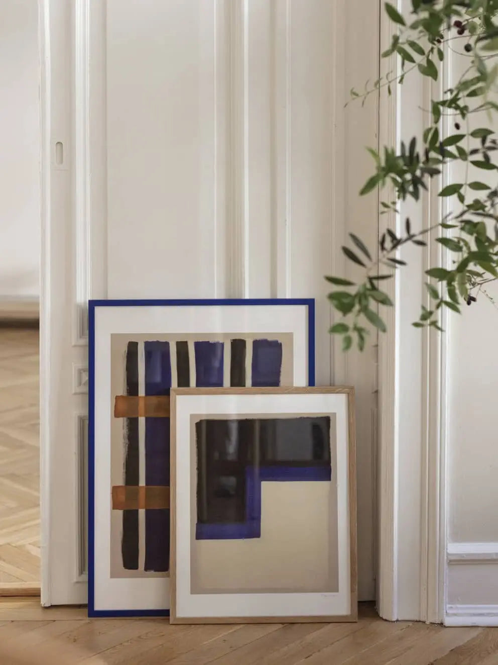 A framed art print, Berit Mogensen Lopez - Checkered (50x70) from The Poster Club, featuring a geometric design, rests against a white wall while a green leafy plant peeks into the scene.