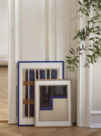 A framed art print, Berit Mogensen Lopez - Checkered (50x70) from The Poster Club, featuring a geometric design, rests against a white wall while a green leafy plant peeks into the scene.