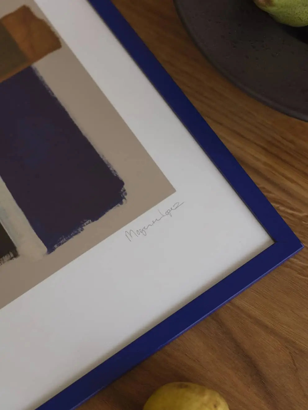 A close-up of The Poster Club Berit Mogensen Lopez - Checkered (50x70) print in a blue frame on wood, with part of a lemon and fruit bowl nearby—evoking the signature style of The Poster Club and artist Berit Mogensen Lopez.