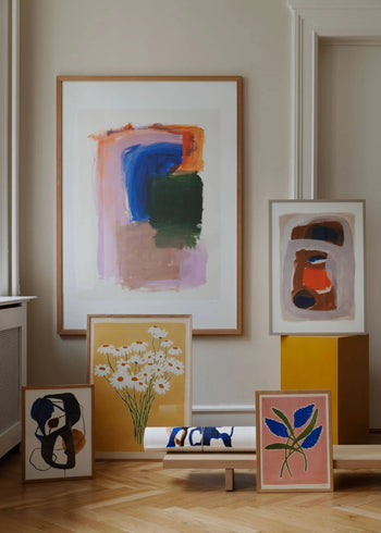 Berit Mogensen Lopez’s Improvise (30x40) from The Poster Club is displayed on a yellow pedestal and floor against a beige wall, its bold brushstrokes and abstract shapes complementing floral-themed art prints.