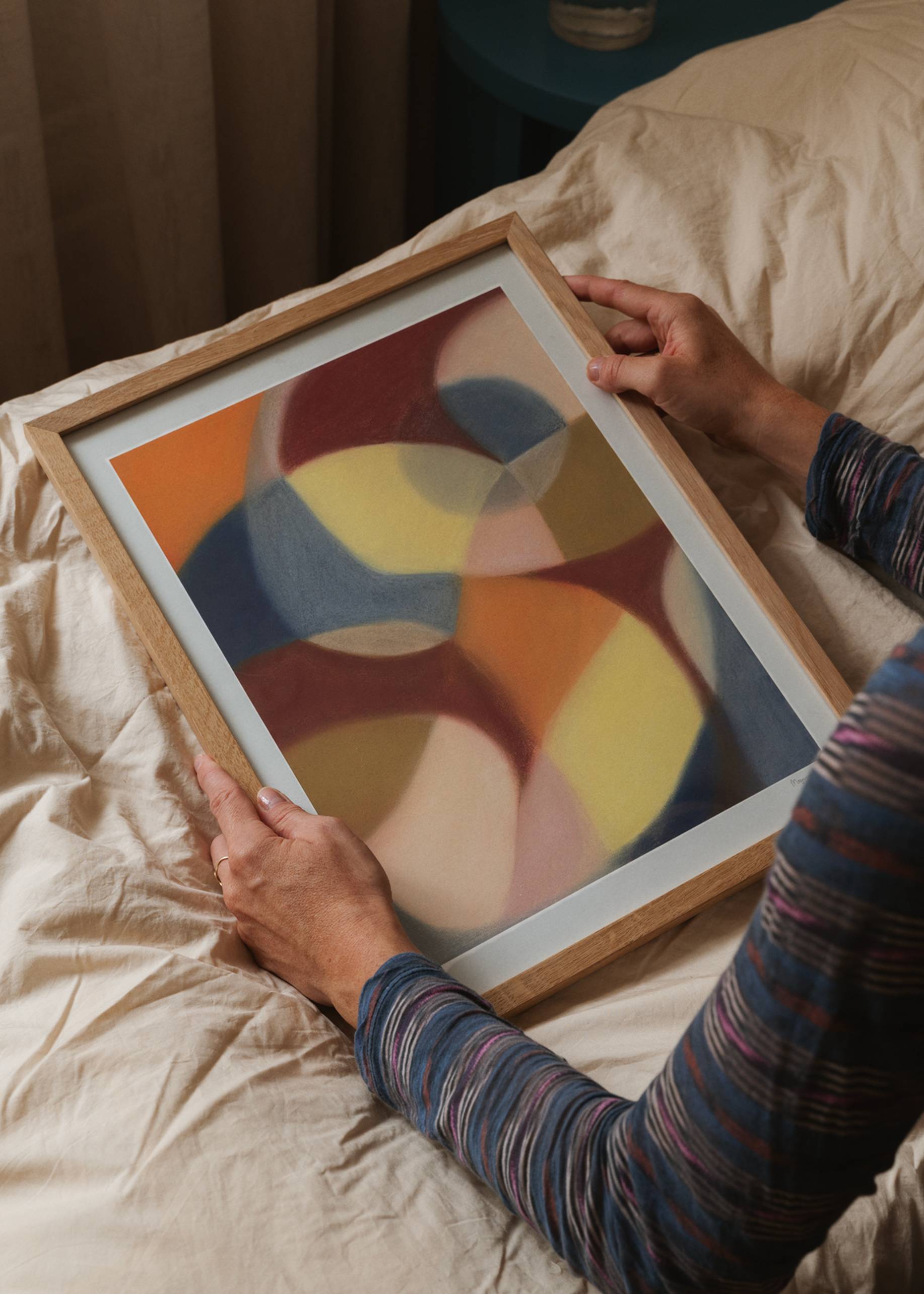 A person sits on a bed with cream sheets, holding The Poster Club Berit Mogensen Lopez - Increase (50x70) print, which displays overlapping shapes in yellow, blue, red, and beige. Available at The Poster Club.