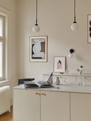 A minimal, light-toned kitchen features a marble countertop with an open book, glass, and water bottle. Two pendant lights hang above while The Poster Club Berit Mogensen Lopez - Levitation (50x70) art print decorates the beige wall.