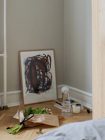 The Poster Club Berit Mogensen Lopez - Checkered (50x70) art print rests against a wall beside books, a carafe, a candle, and a small lamp; a paper-wrapped bouquet of flowers lies on the floor. Available at The Poster Club.