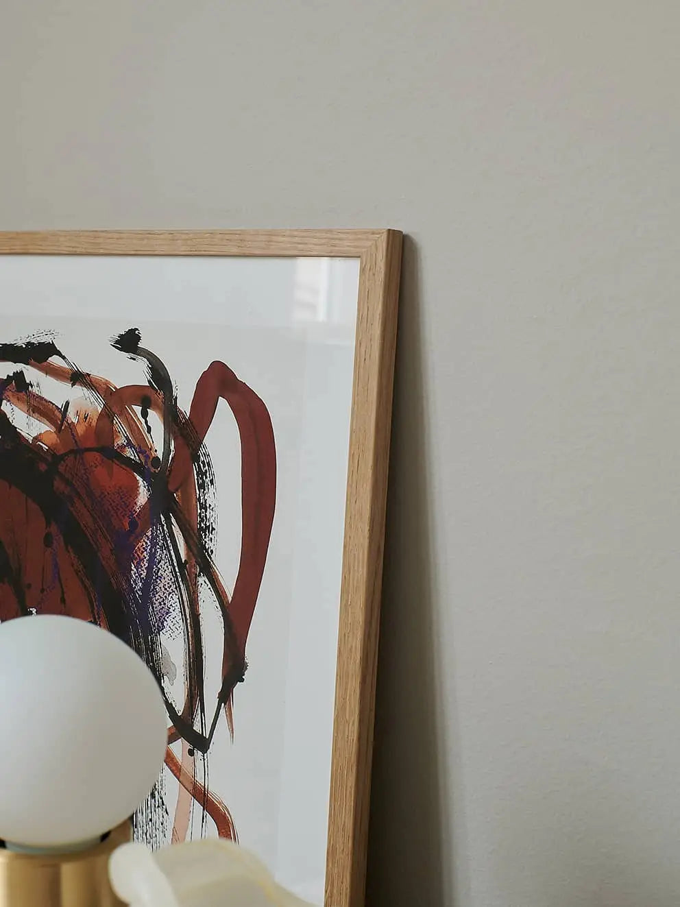 A close-up of The Poster Club Berit Mogensen Lopez - Mixed Media (50x70) art print with striking red and black brushstrokes, framed and leaning against a beige wall, with part of a round white lamp in the foreground.