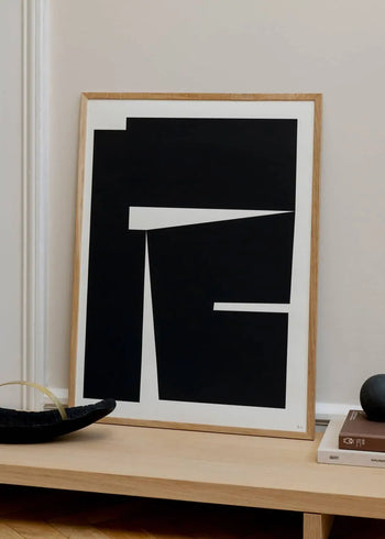 The Poster Club Bycdesign Studio - Composition 04 (50x70) art print displays bold black geometric shapes on white, framed in light wood and set against a beige wall on a light wooden surface.