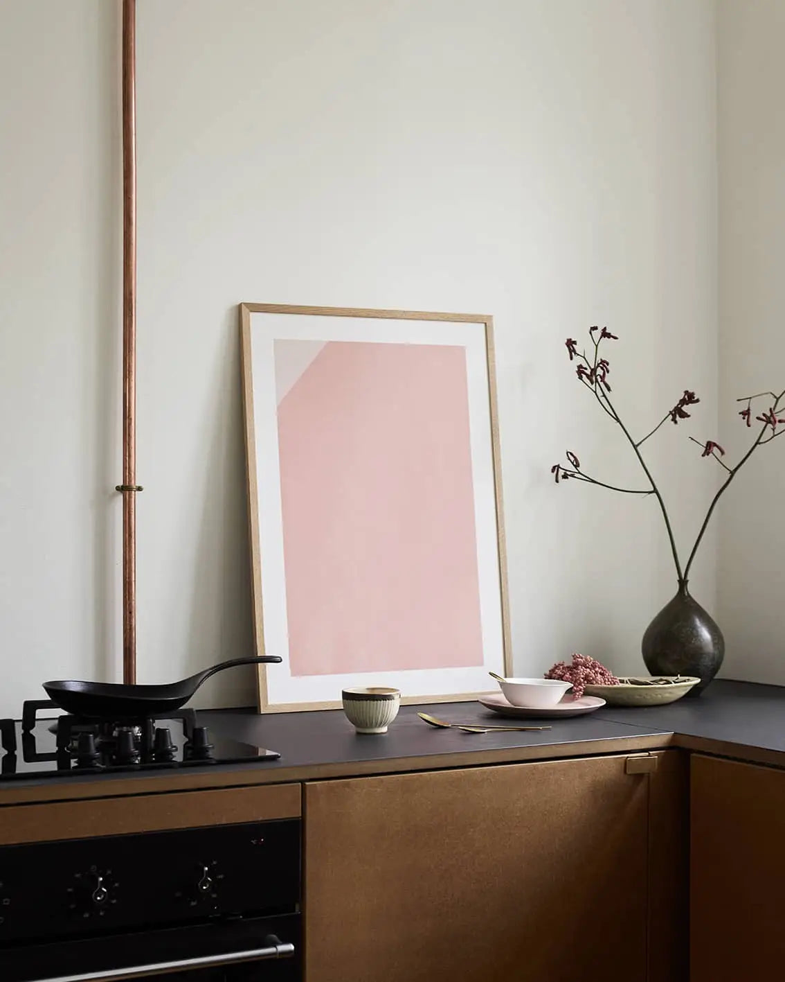 Minimalist kitchen corner with The Poster Clubs Jaguar 01 (50x70) art print by Anastasia Krasilnikova, a black countertop, framed abstract pink artwork, dark vase with branches, frying pan on the stove, and warm neutral tones.