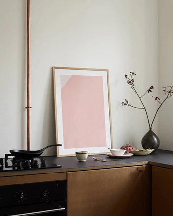 Minimalist kitchen corner with The Poster Clubs Jaguar 01 (50x70) art print by Anastasia Krasilnikova, a black countertop, framed abstract pink artwork, dark vase with branches, frying pan on the stove, and warm neutral tones.