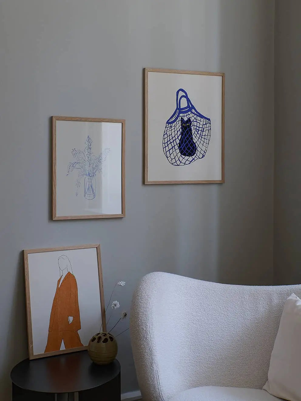 A cozy corner with a white textured armchair, a round black side table with a vase of dried flowers, and The Poster Club’s Chloe Purpero Johnson - The Cat’s In The Bag (30x40) fine art giclée print on light gray walls.