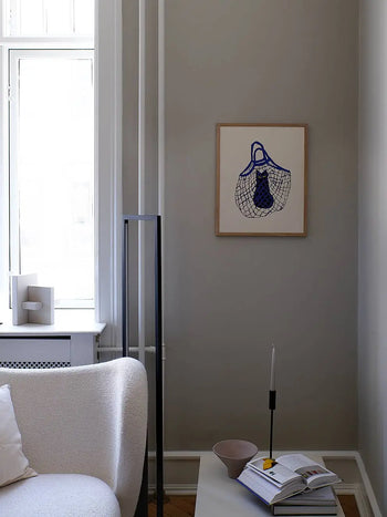 A minimalist living room showcases a white textured sofa, a side table with an open book, bowl, and candle, and The Poster Club Anna Mörner - Statue (50x70) art print hanging on the light gray wall near a tall window.