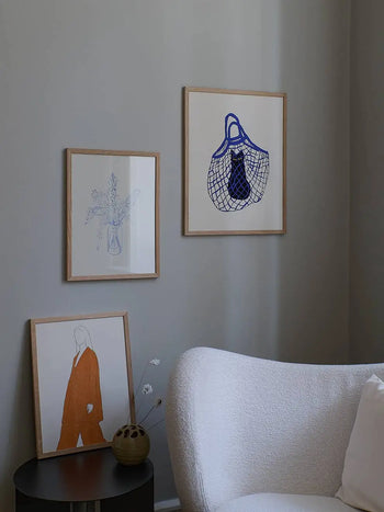 A cozy room corner with a white textured armchair, a small black round table, and The Poster Club’s Chloe Purpero Johnson - The Cat’s In The Bag (50x70) giclée print on the gray wall.