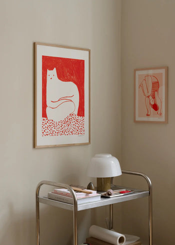 A minimalistic corner features The Poster Club Cinzia Franceschini - Cat In Red (50x70) giclée print, paired with an abstract animal artwork. Below, a metal cart holds a white lamp, books, and papers.