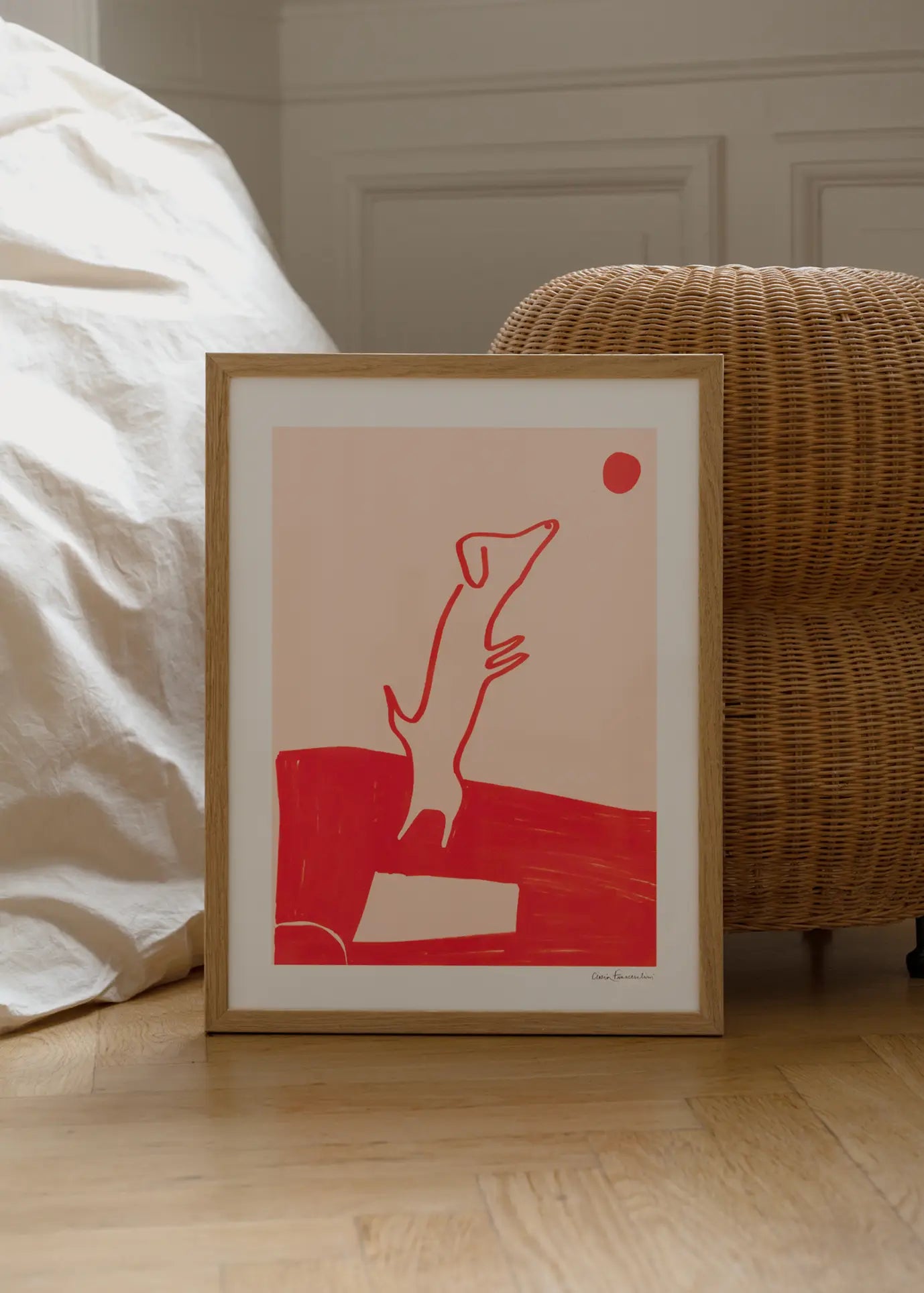 The Poster Club’s Cinzia Franceschini - Divertissement (50x70) is a framed fine art giclée print featuring a red minimalist dog on its hind legs reaching for a red ball, displayed by a woven chair and white fabric on a wooden floor.
