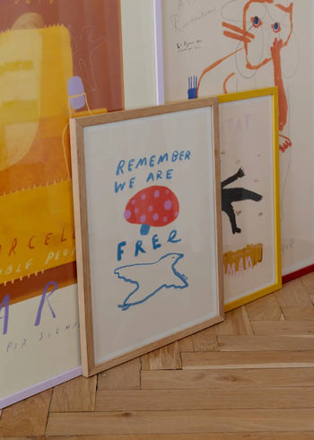 Three framed art prints rest on a wooden floor. The central Sarah Williams print reads REMEMBER WE ARE FREE with a red shape, blue text, and a white bird; the others feature abstract drawings and The Poster Club’s Das Rotes Rabbit - Afternoon Communiqué.