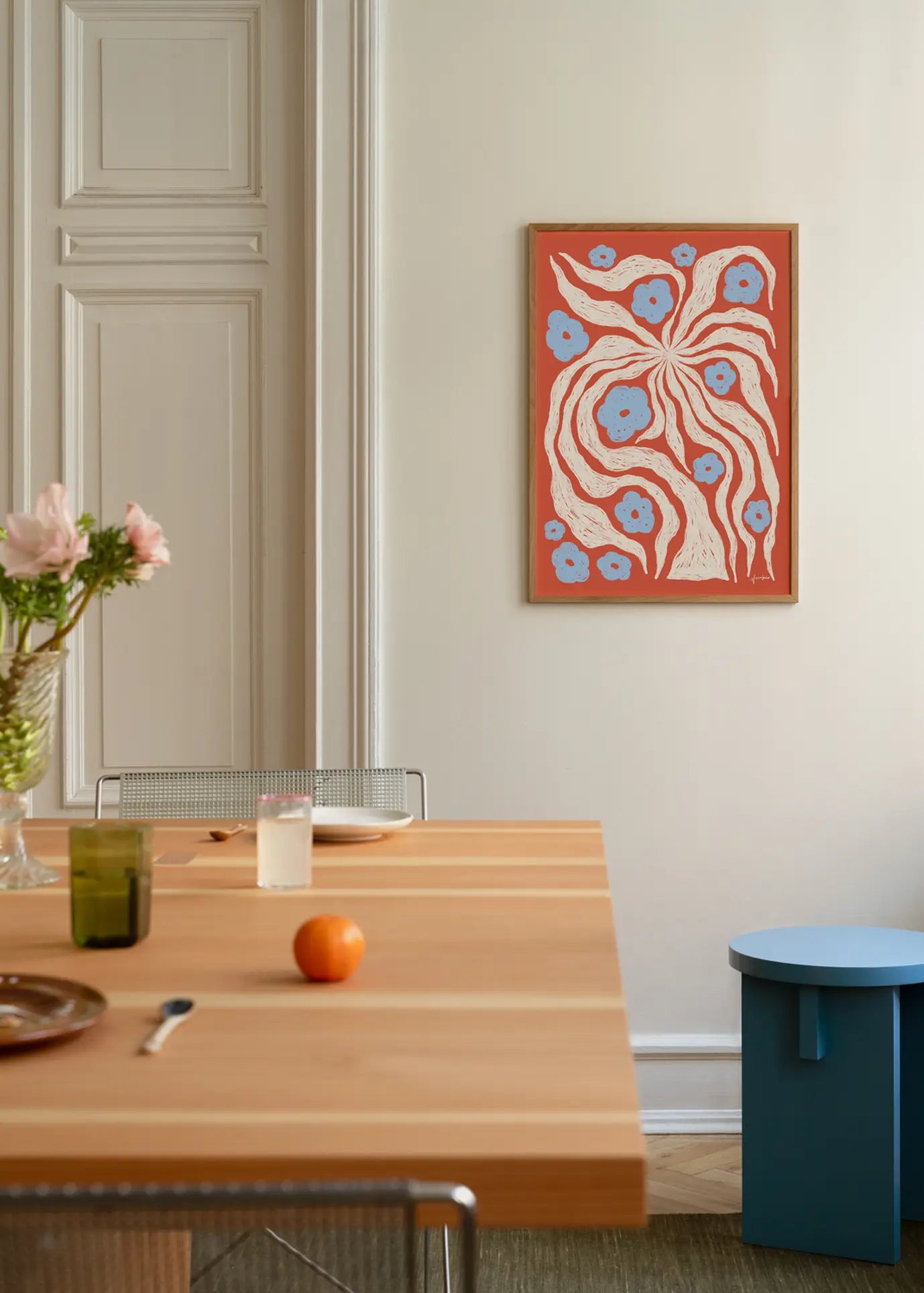 A modern dining area features a light wood table with assorted dishes, glasses, pink flowers, and an orange. On the wall above a small blue round table hangs The Poster Club Hanna KL - Misty (30x40) art print in red, white, and blue.