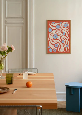 A minimalist dining area with a light wooden table, wire chair, and blue side table features The Poster Club Hanna KL - Misty (50x70) artwork above flowers, glasses, a plate, and an orange.