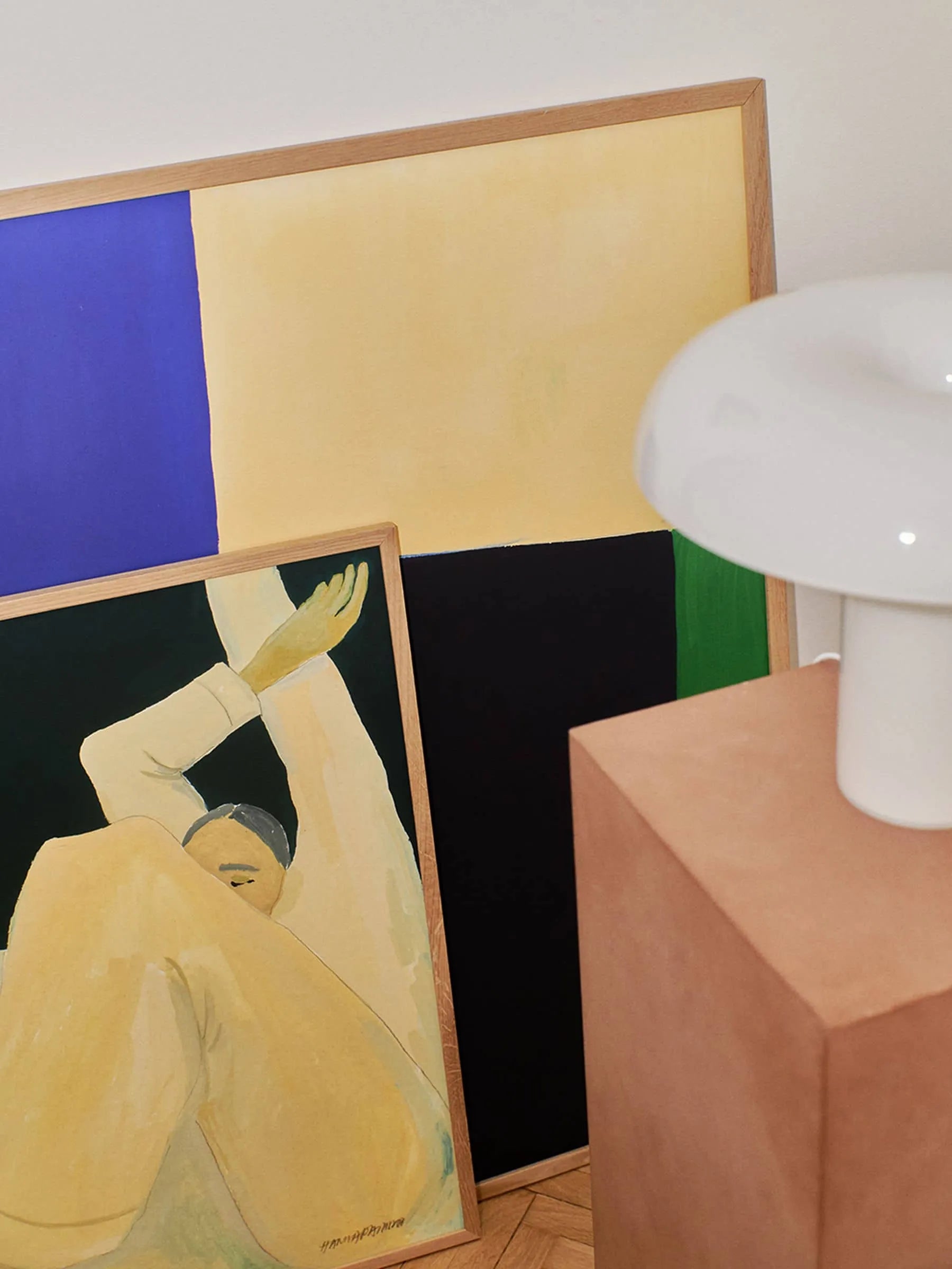 Hanna Petersons Court (30x40) fine art giclée print from The Poster Club leans against the wall, accompanied by an abstract piece. In the foreground, a white lamp sits atop a peach pedestal.