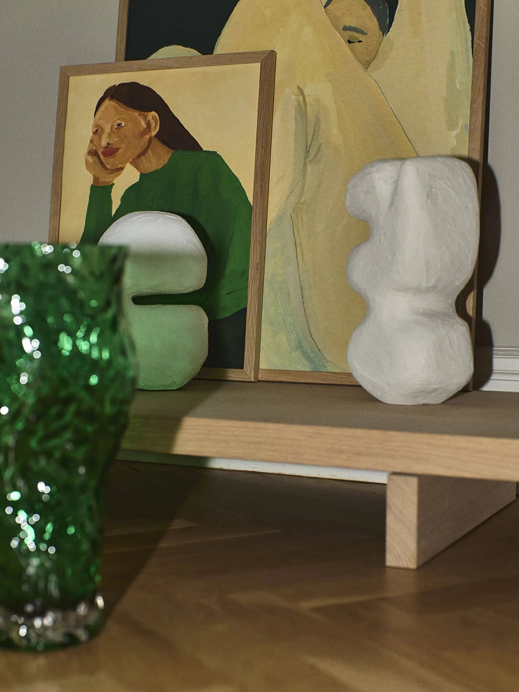 A Green Guise (30x40) by Hanna Peterson for The Poster Club is displayed on a light wooden shelf with two abstract white sculptures and two Daydreamer Collection giclée prints against a neutral wall.