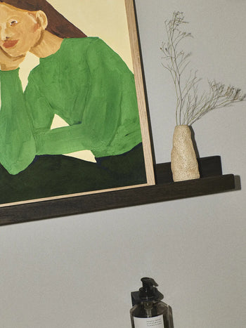 The Poster Clubs Hanna Peterson - Green Guise (50x70) print, from The Daydreamer Collection, is framed and displayed on a dark shelf beside a beige vase with dried foliage and a black soap dispenser against a light wall.