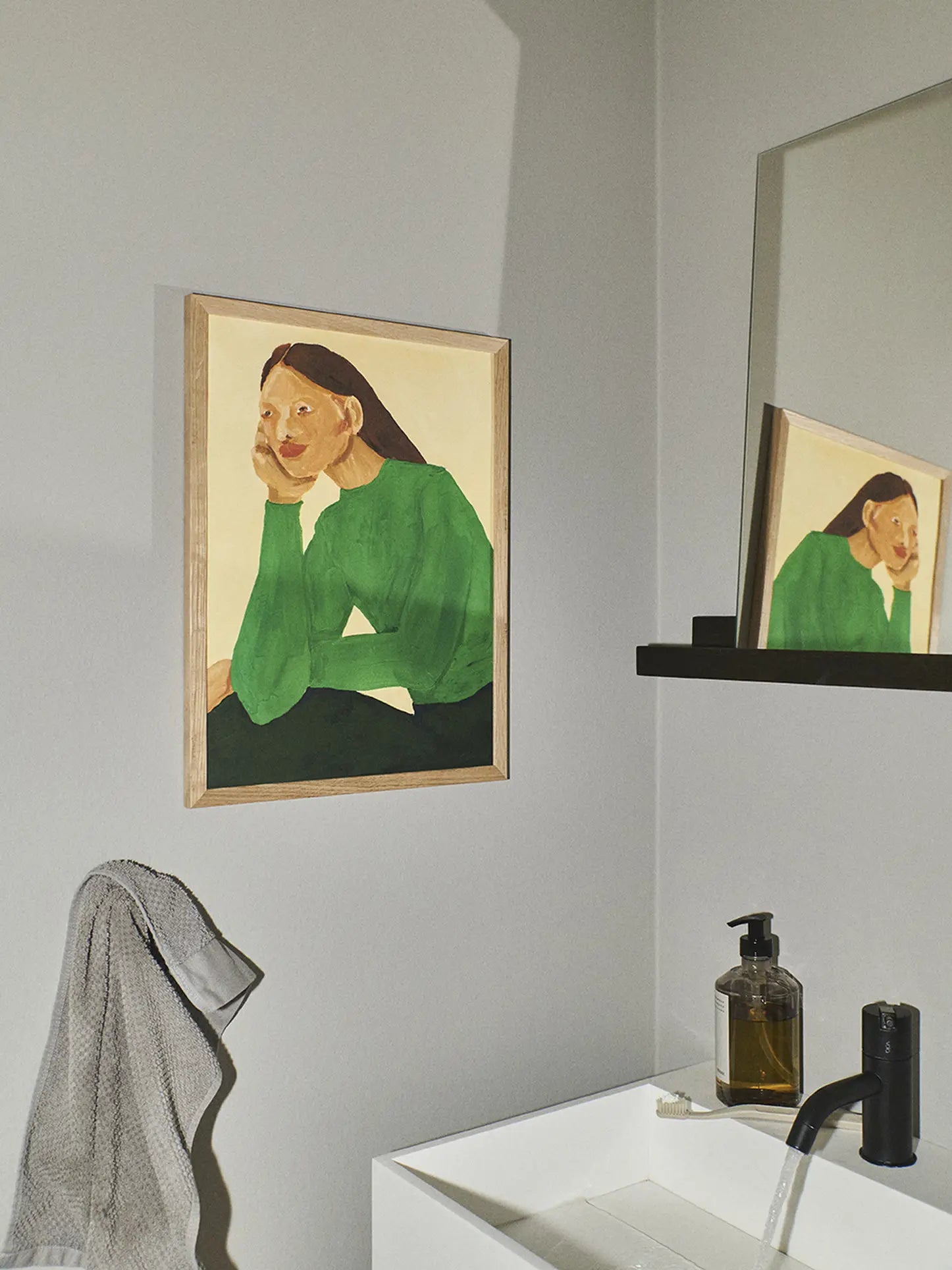 A minimalist bathroom showcases The Poster Club’s Hanna Peterson - Green Guise (50x70) print, depicting a woman in a green top, mounted above the sink and near a towel, with the mirror reflecting the artwork.