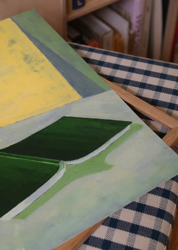 A close-up of The Poster Club Hanna Peterson - Green Guise (50x70) fine art giclée print, featuring green and yellow abstract shapes on blue and white checkered fabric, with a wooden frame and bookshelf in the background.