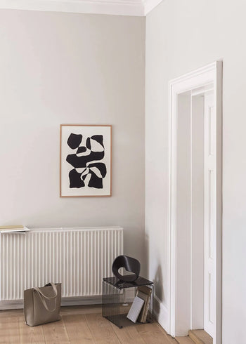 A minimalist room with light gray walls features the Hanna Peterson - Impossible Island (30x40) Giclée print by The Poster Club above a white radiator, alongside a beige bag, black chair, and small side table near a white door and light wood floor.