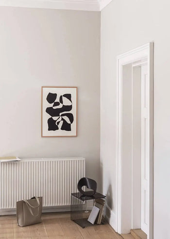 A minimalist room with light gray walls features The Poster Club Hanna Peterson - Impossible Island (50x70) giclée print above a radiator, a black modern chair, side table, tote bag, and books on a light wooden floor.