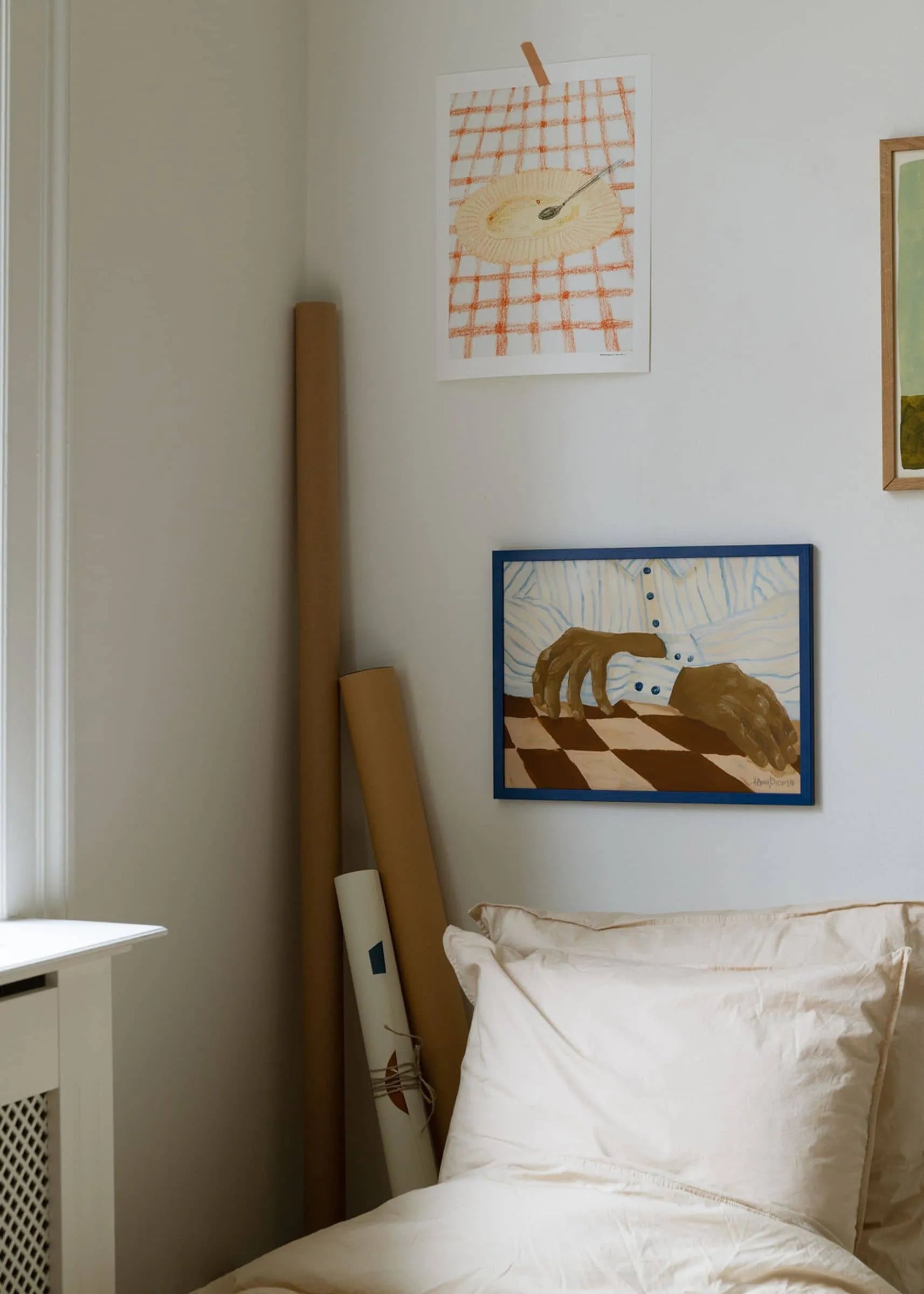 A cozy corner with a bed topped with cream pillows, Hanna Petersons Light Stripes (30x40) by The Poster Club on the wall, and rolled posters from The Poster Club leaning nearby in soft natural light.