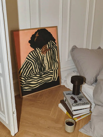 The Poster Club’s Ana Frois - Orange Pendant (50x70) art print rests against the wall by a bed, with books, a lamp, and a coffee cup arranged on the wooden floor.