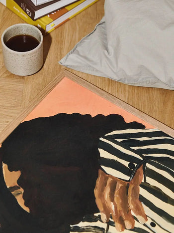 The Poster Club Anna Johansson - Monday (30x40) art print rests on a wooden floor, beside a coffee cup, books, and a light gray cushion, reflecting the curated elegance of The Poster Club’s signature aesthetic.
