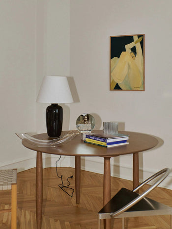 A round wooden table with a black lamp, books, and decor sits on a herringbone wood floor surrounded by chairs. On the cream wall hangs The Dream (30x40) giclée print by Hanna Peterson from The Poster Club.