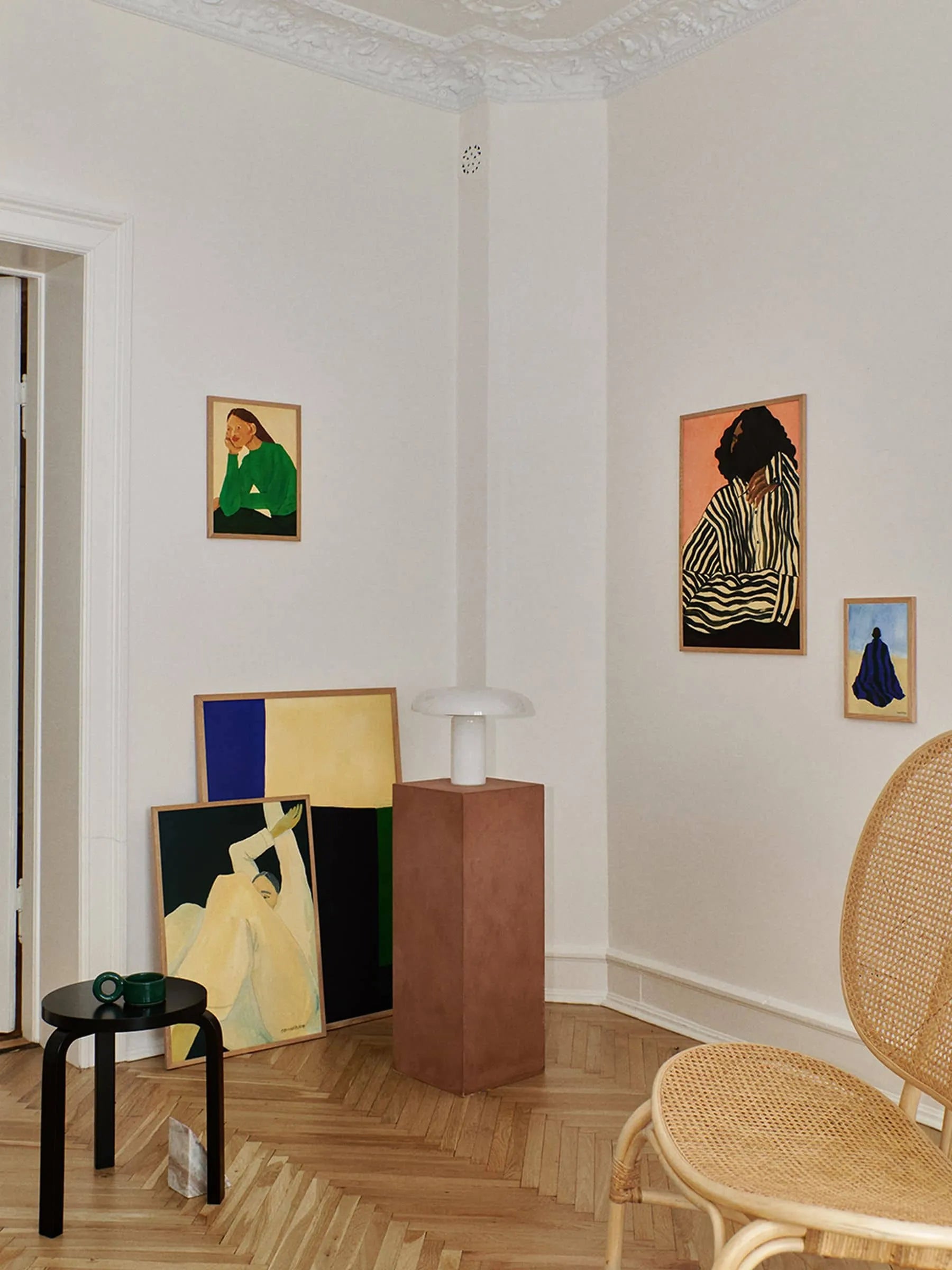A minimalist room with light walls, modern art, a Fine art giclée print—The Dream (30x40) by Hanna Peterson from The Poster Club—a round wicker chair, small black table with green cup, and a white lamp on a brown pedestal.