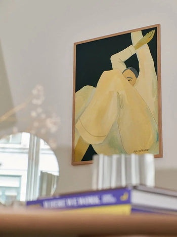 The Poster Clubs Hanna Peterson - The Dream (50x70) framed art print features a person in loose yellow clothing with raised arms, plus a mirror, blurred glass, and stacked books on a table in the foreground.