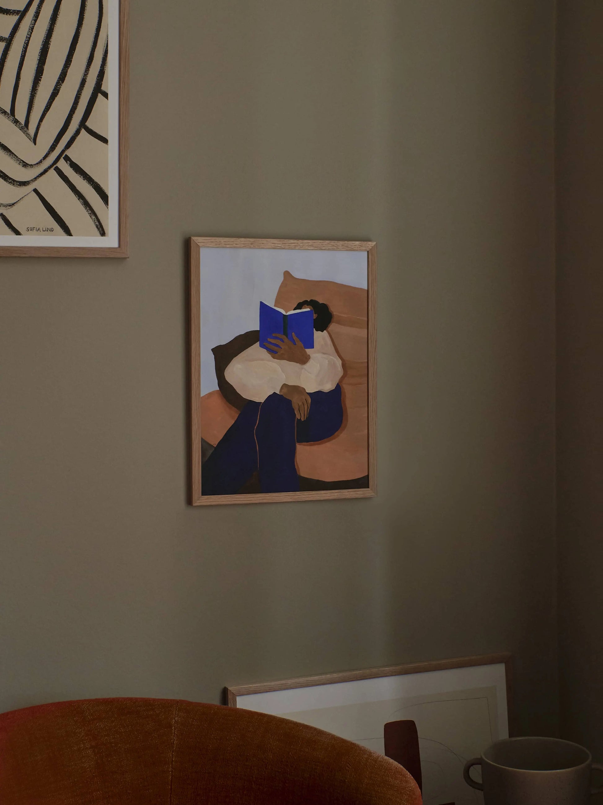 A framed giclée print, The Togo (30x40) by Hanna Peterson from The Poster Club, hangs on a taupe wall. It depicts a person reading a blue book on a couch in a cozy, softly lit setting with more artwork and a cup nearby.