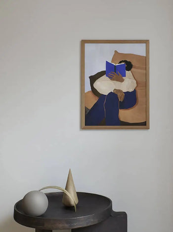 The Poster Club Hanna Peterson - The Togo (30x40) is a giclée print on premium art paper, depicting a person reading a blue book with a round vase and geometric sculpture on a dark table, perfect for minimalist, modern decor.