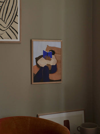 The Poster Club Hanna Peterson - The Togo (50x70) is a framed fine art giclée print showing a person reading on a couch. Created with warm, muted colors and archival materials from The Poster Club, it exudes a cozy, timeless feel.