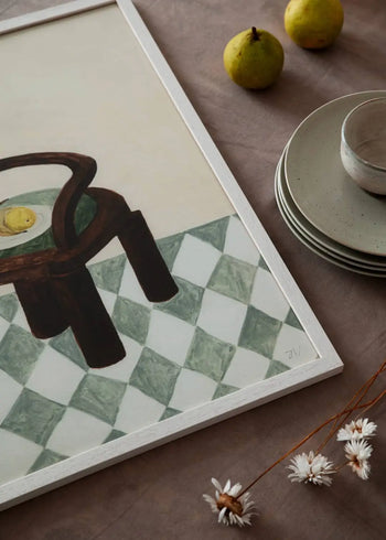 The Poster Clubs Berit Mogensen Lopez - Checkered (50x70) art print depicts a chair on a green and white checkered floor, displayed on a taupe surface beside stacked ceramic plates, a cup, two green pears, and dried white flowers.