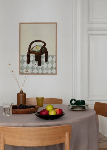 A round table with a beige tablecloth holds fruit, drinks, and green cups. Wooden chairs surround it; “Chair with Fruit” by Isabelle Vandeplassche (30x40) from The Poster Club decorates the white wall.