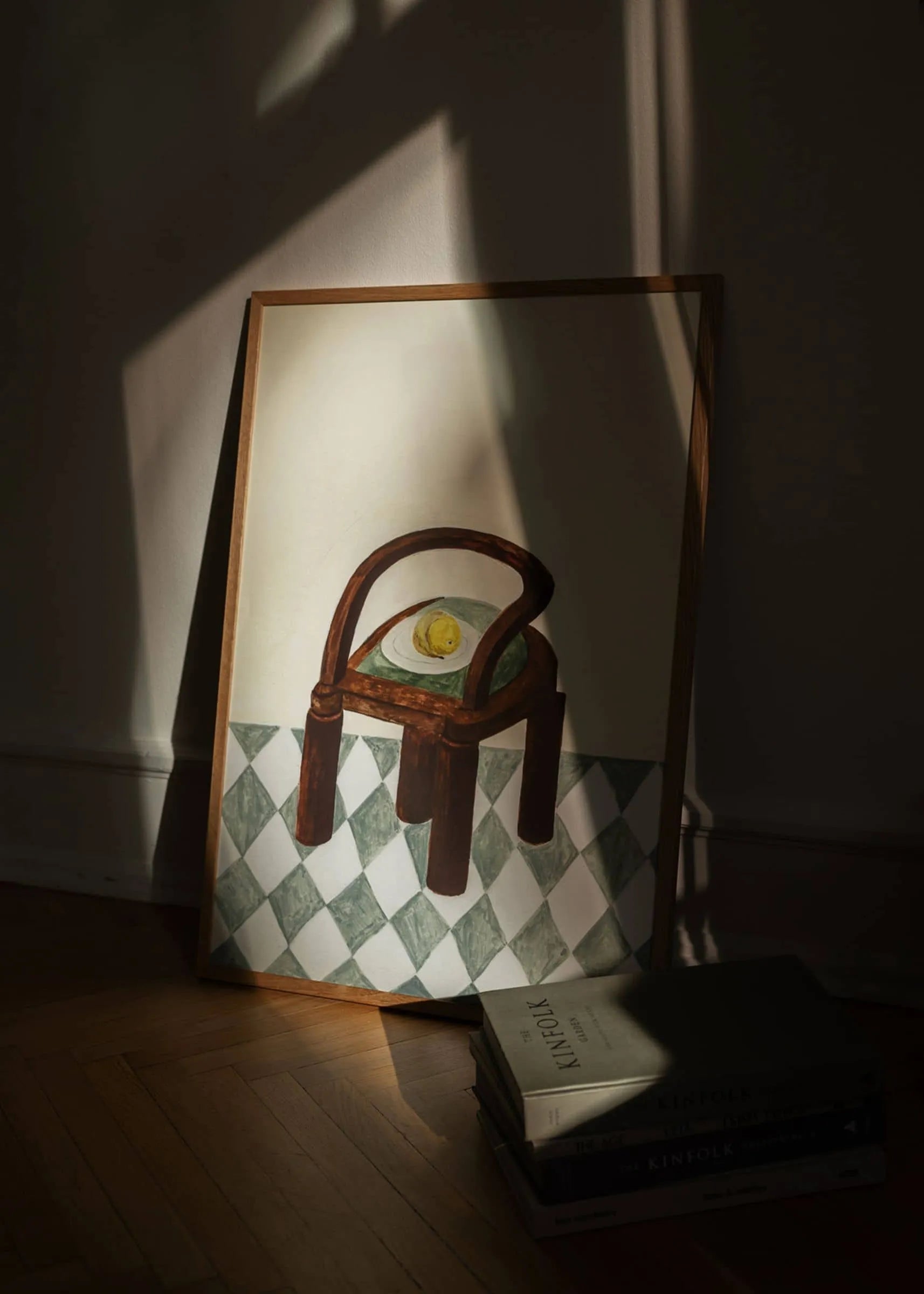 The Poster Club’s Isabelle Vandeplassche - Chair with Fruit (30x40) art print shows a wooden chair with a lemon on its seat beside books on the floor, all set against a sunlit wall and casting gentle shadows.