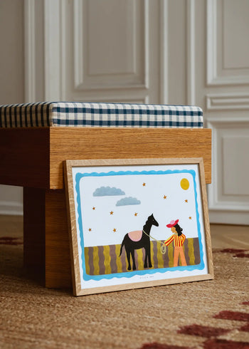 The Poster Club Kelly Jay - A Journey Begins (50x70) art print, inspired by Kelly Jay, features a person with a rope and black horse under a bright sky, framed and displayed against a bench in a stylish room with white paneled walls.