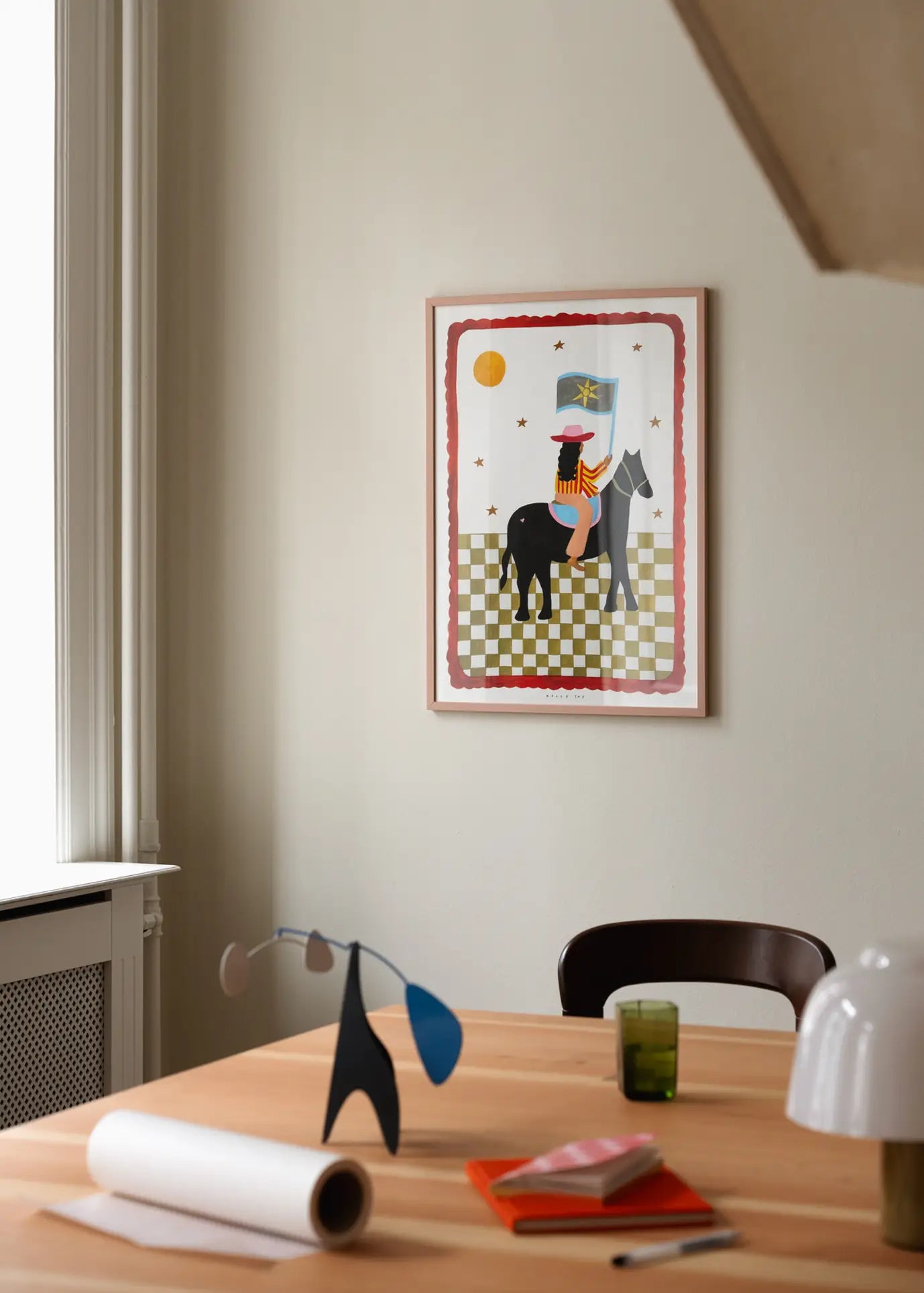A framed The Poster Club Kelly Jay - The More I Travel (30x40) art print hangs on a light wall above a wooden table with a mobile, rolled paper, green glass, red notebook, and white lamp. Natural light comes in from the left.