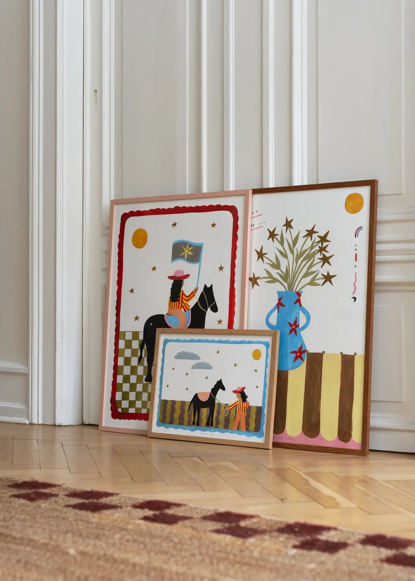Three vibrant framed art prints from The Poster Club rest on a wooden floor; two show a figure on horseback with the sun, and the third is Kelly Jays The More I Travel (30x40), depicting a blue vase with star-shaped flowers.