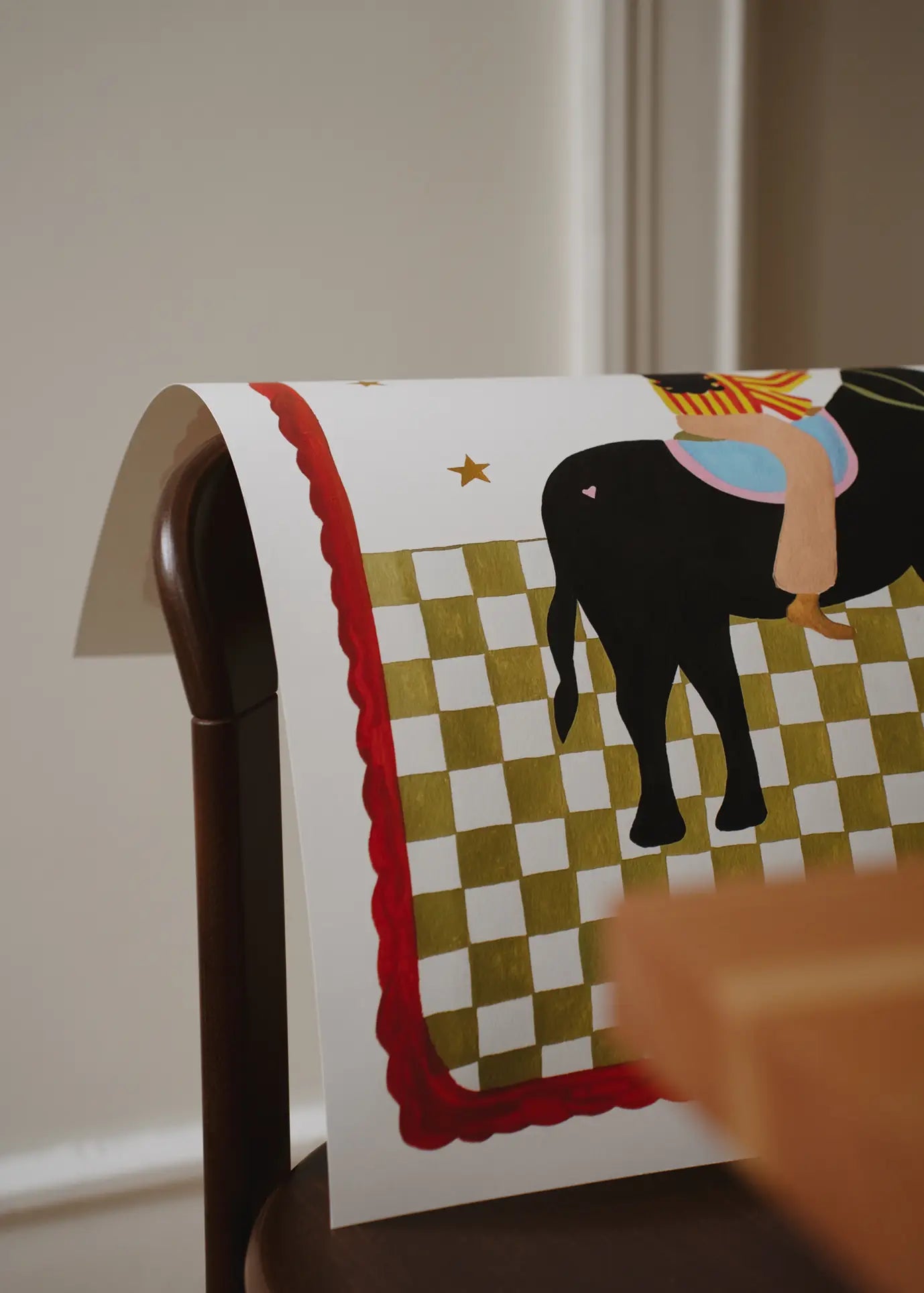 The Poster Club Kelly Jay - The More I Travel (30x40) art print features a checkered design, red border, and part of a black saddled horse over a brown chair near a wooden table. Artwork by Kelly Jay from The Poster Club.