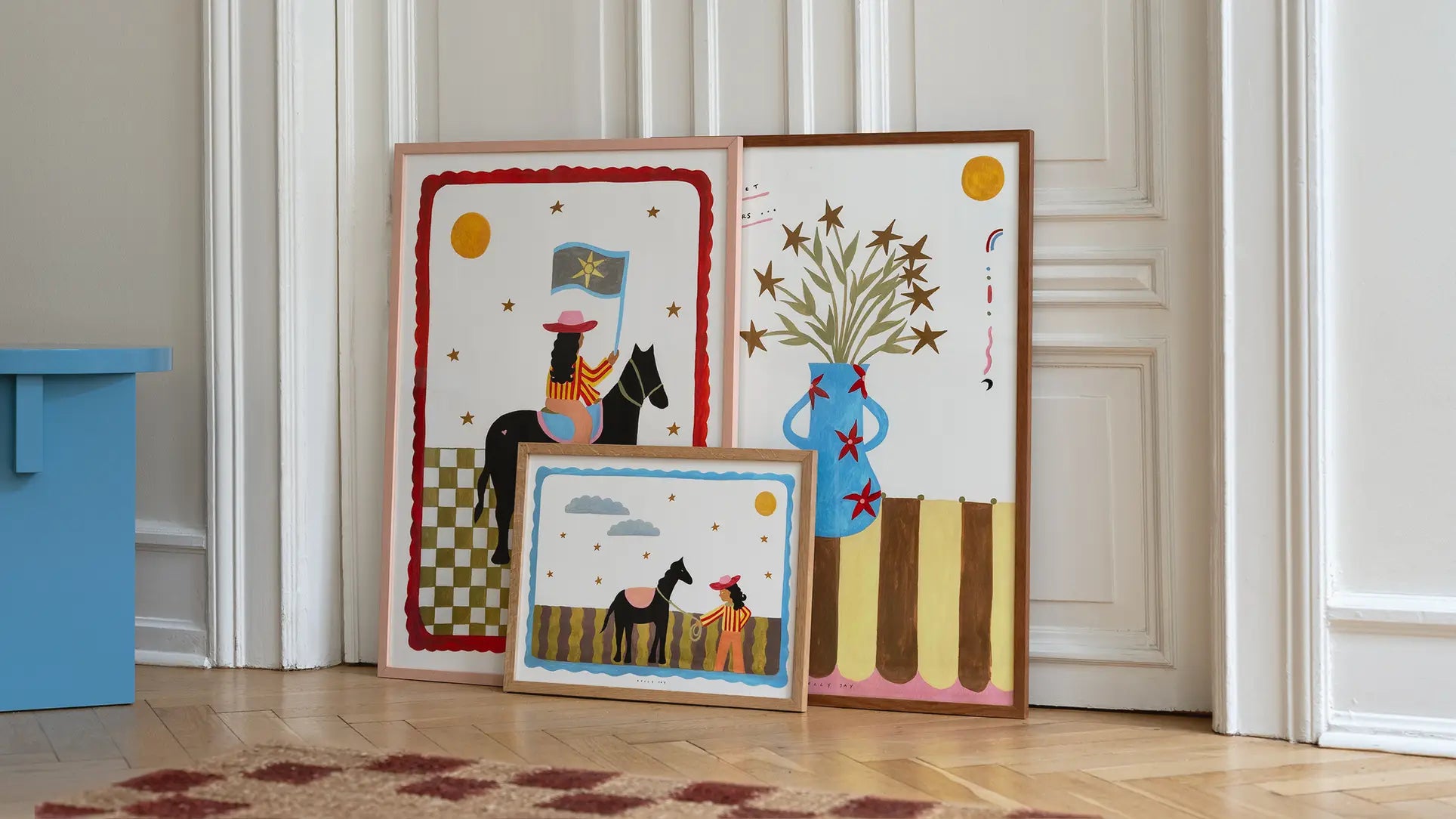 The Poster Club Kelly Jay - The More I Travel (30x40) art print rests against a white paneled wall on a wooden floor, showcasing bold shapes and warm colors in stylized figures, horses, and a vase. A patterned rug is partially visible.