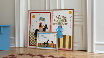 The Poster Club Kelly Jay - The More I Travel (30x40) art print rests against a white paneled wall on a wooden floor, showcasing bold shapes and warm colors in stylized figures, horses, and a vase. A patterned rug is partially visible.