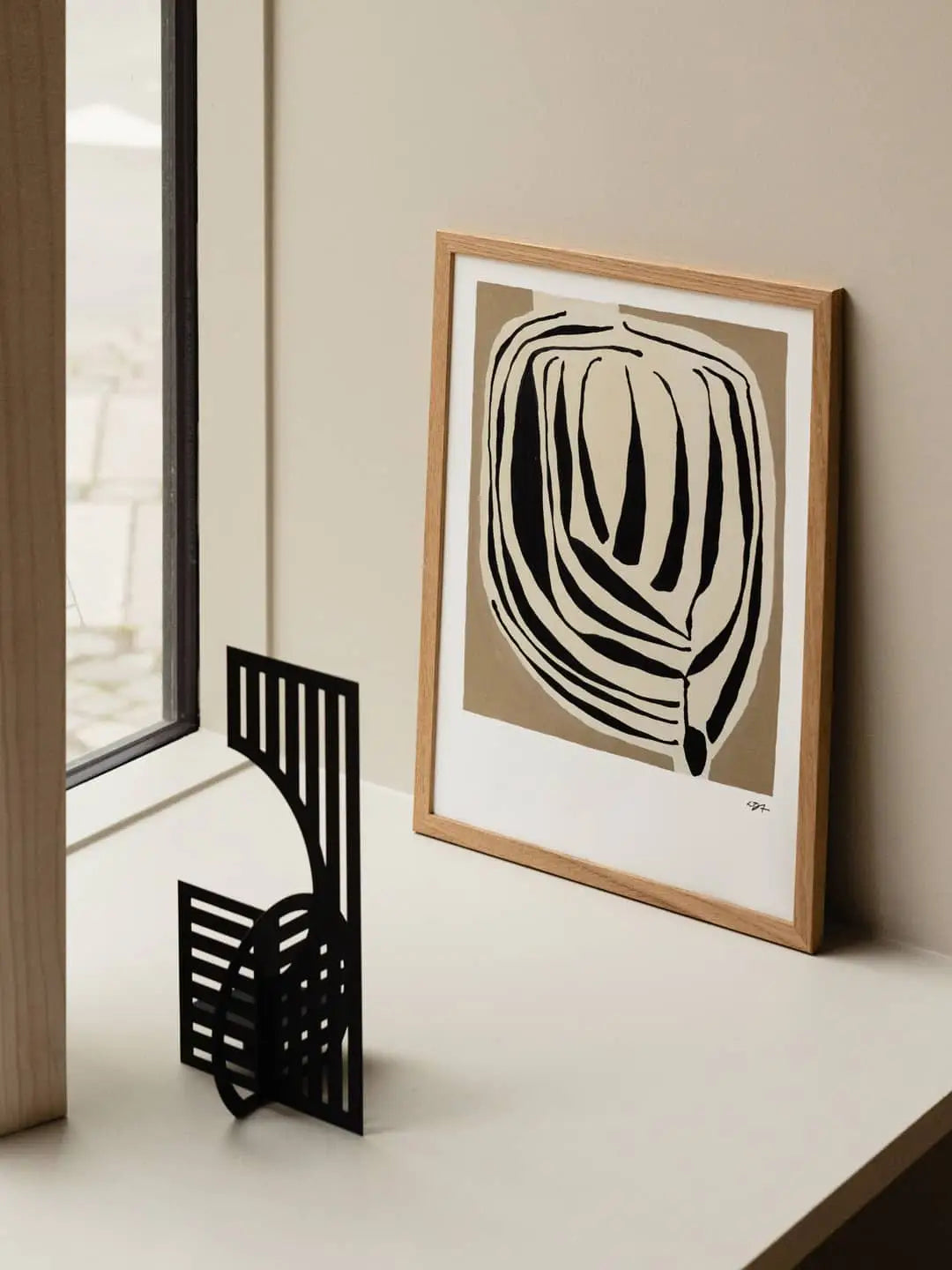 The Poster Club Leise Dich Abrahamsen - Layered (50x70) framed print leans against a beige wall on a windowsill, while a small black geometric metal sculpture sits in front, adding modern flair.